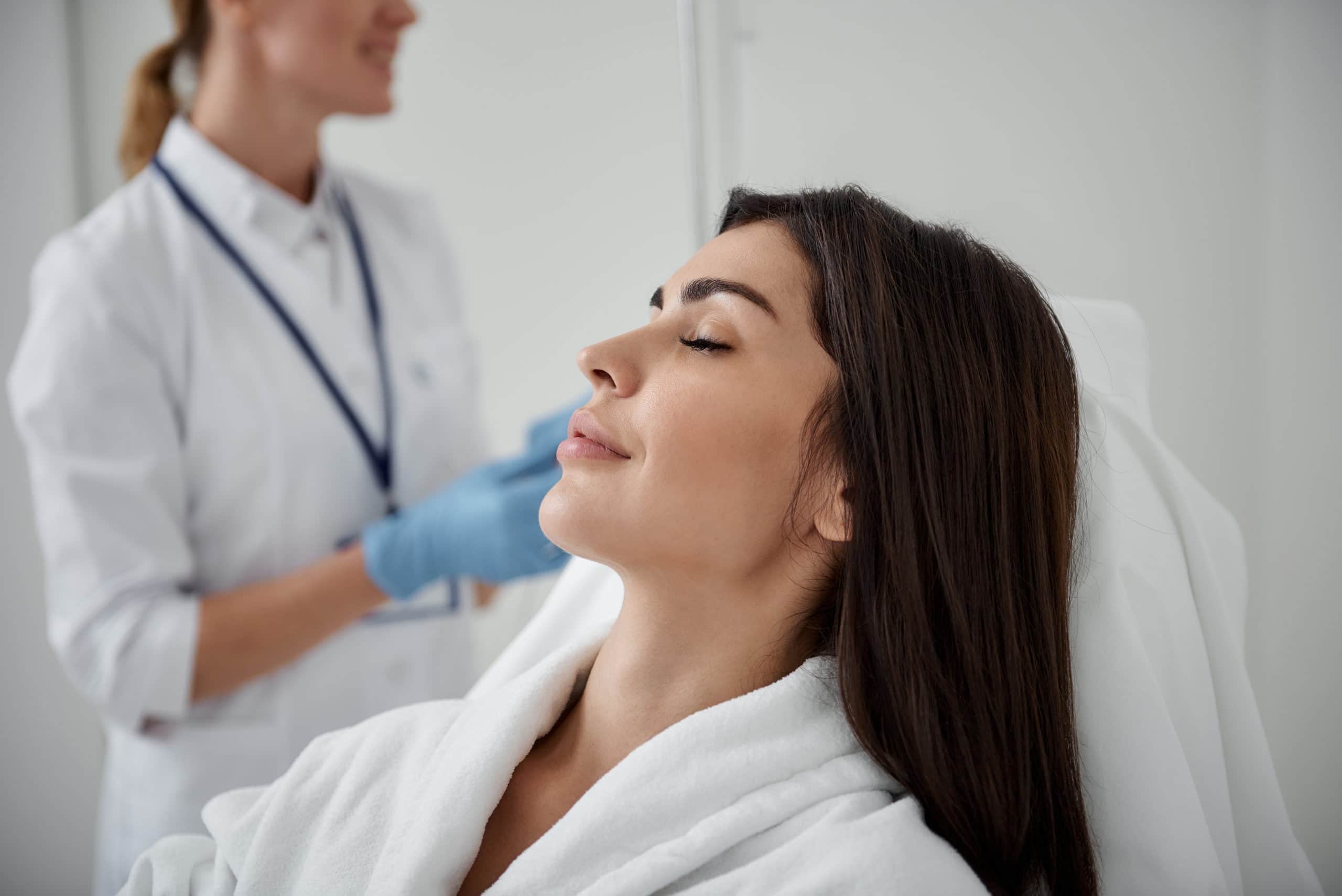 Beautiful lady resting while doctor checking intravenous drip Side view portrait of attractive woman in white bathrobe lying with closed eyes during medical procedure
